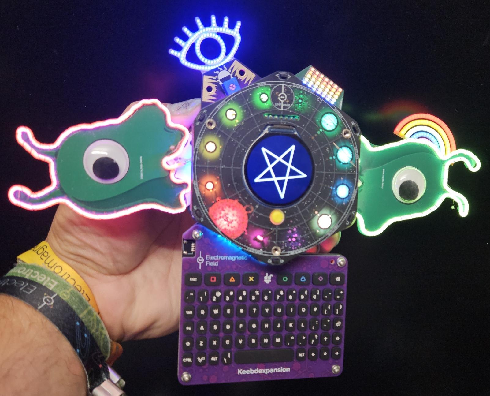 An EMF Camp spaceagon with keyboard attached, bender had hexpansions left and right, right one with a rainbow hat, top left is a glowing blue illuminati eye, top right is sorta kinda pride flag LED rectangule.